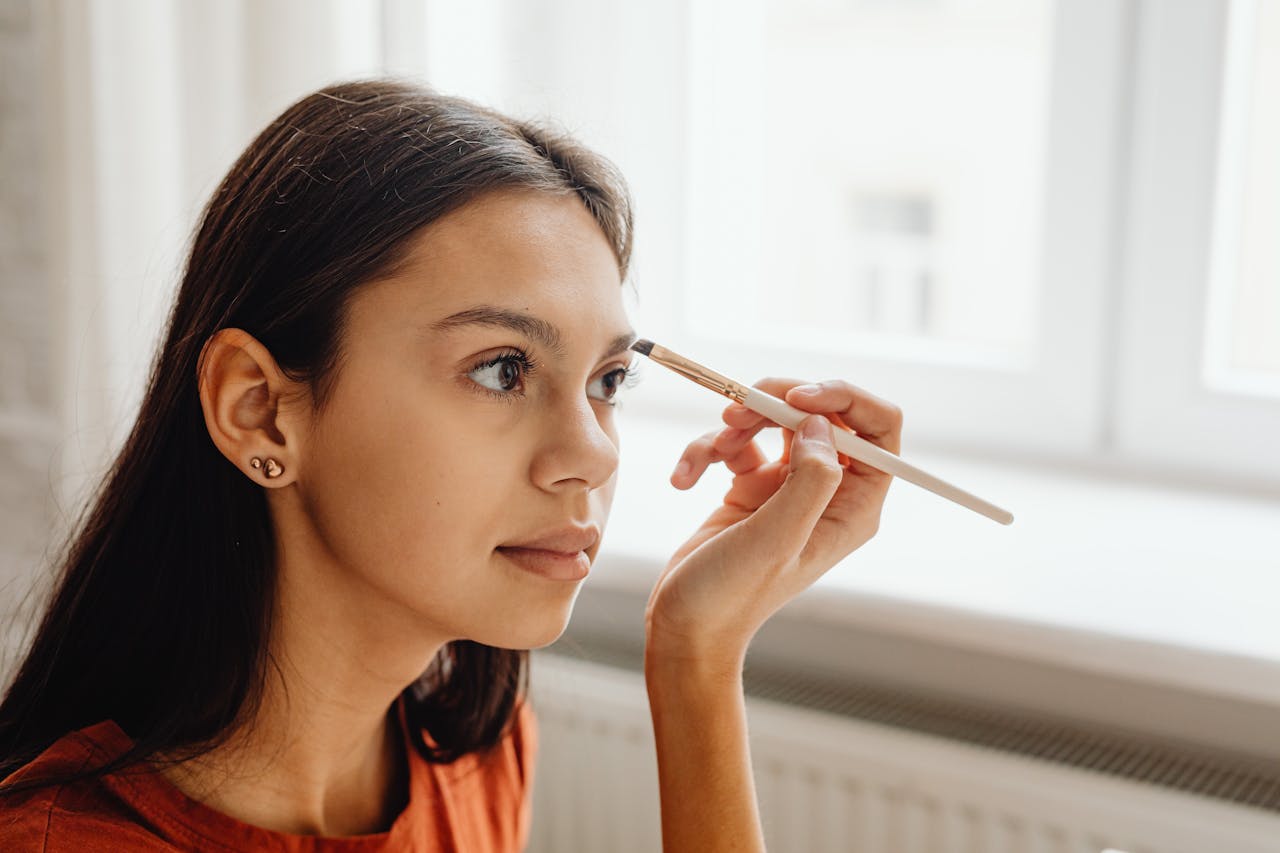 A young woman skillfully applying makeup with a brush by a window indoors.